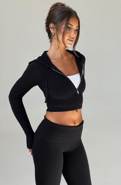 Sudadera negra de punto con capucha y cremallera, vista lateral con bajo ajustado acanalado.